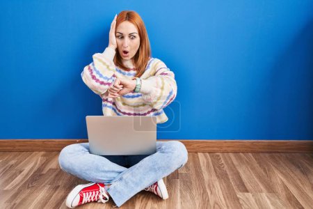 Téléchargez les photos : Jeune femme utilisant un ordinateur portable à la maison assis sur le sol en regardant l'heure de la montre inquiet, peur de se faire tard - en image libre de droit