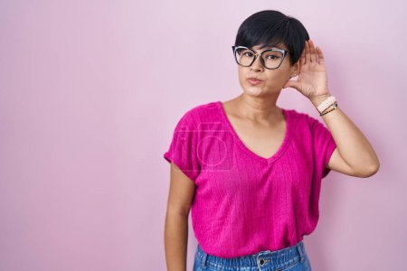 Téléchargez les photos : Jeune femme asiatique aux cheveux courts debout sur fond rose souriant avec la main sur l'oreille en écoutant une rumeur ou des commérages. concept de surdité. - en image libre de droit