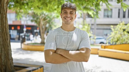 Photo for Cheerful young hispanic man exuding happiness and confidence, smiling and laughing while making a crossed arms gesture, standing tall in a vibrant, green park - Royalty Free Image