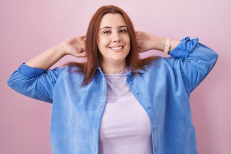 Téléchargez les photos : Jeune femme hispanique aux cheveux roux debout sur fond rose relaxant et étirant, bras et mains derrière la tête et le cou souriant heureux - en image libre de droit
