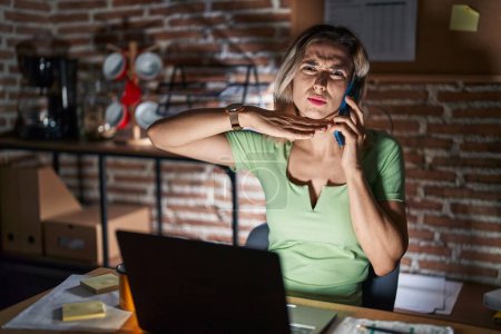 Téléchargez les photos : Jeune belle femme travaillant au bureau la nuit parlant au téléphone tranchant la gorge avec la main comme un couteau, menace d'agression avec une violence furieuse - en image libre de droit