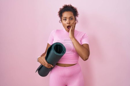 Photo for Young hispanic woman with curly hair holding yoga mat over pink background afraid and shocked, surprise and amazed expression with hands on face - Royalty Free Image