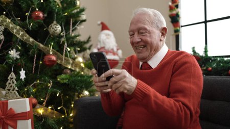 Photo for Senior using smartphone sitting by christmas tree at home - Royalty Free Image