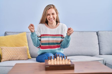 Photo for Young blonde woman playing chess sitting on the sofa screaming proud, celebrating victory and success very excited with raised arm - Royalty Free Image