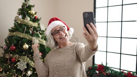 Foto de Mujer de mediana edad con el pelo gris que tiene videollamada de pie junto al árbol de Navidad en casa - Imagen libre de derechos