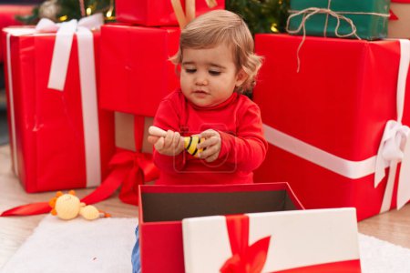 Foto de Adorable niño hispano desempacando regalo sentado junto al árbol de Navidad en casa - Imagen libre de derechos
