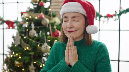 Téléchargez les photos : Femme hispanique mature avec les cheveux gris priant assis près de l'arbre de Noël à la maison - en image libre de droit