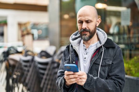 Téléchargez les photos : Jeune homme chauve utilisant un smartphone avec une expression sérieuse dans la rue - en image libre de droit