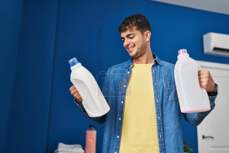 Photo for Young hispanic man washing clothes holding detergent bottles at laundry room - Royalty Free Image