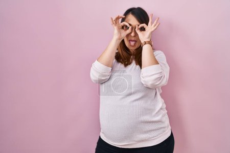 Photo for Pregnant woman standing over pink background doing ok gesture like binoculars sticking tongue out, eyes looking through fingers. crazy expression. - Royalty Free Image