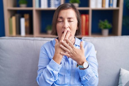 Foto de Mujer hispana de mediana edad sentada en el sofá en casa aburrida bostezando cansada cubriendo la boca con la mano. inquietud y somnolencia. - Imagen libre de derechos
