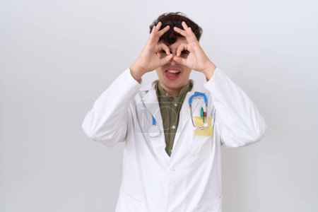 Photo for Young non binary man wearing doctor uniform and stethoscope doing ok gesture like binoculars sticking tongue out, eyes looking through fingers. crazy expression. - Royalty Free Image