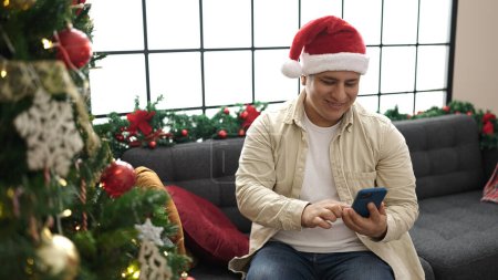 Foto de Joven hombre hispano usando smartphone celebrando la Navidad en casa - Imagen libre de derechos