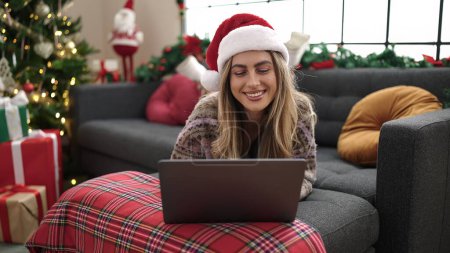 Foto de Mujer rubia joven usando el ordenador portátil tumbado en el sofá por el árbol de Navidad en casa - Imagen libre de derechos