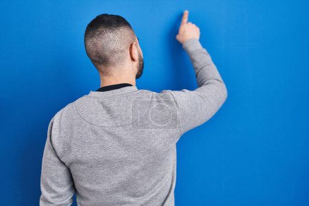 Foto de Hombre hispano de pie sobre fondo azul posando hacia atrás apuntando hacia adelante con la mano del dedo - Imagen libre de derechos