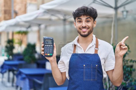 Téléchargez les photos : Homme arabe avec barbe portant tablier de serveur à la terrasse du restaurant tenant dataphone souriant heureux pointant avec la main et le doigt sur le côté - en image libre de droit