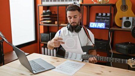 Téléchargez les photos : Jeune homme arabe musicien jouant de la guitare électrique composer chanson à l'aide d'un ordinateur portable au studio de musique - en image libre de droit