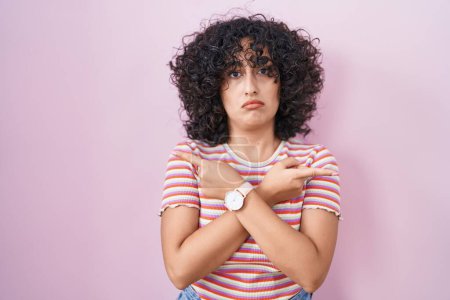 Photo for Young middle east woman standing over pink background pointing to both sides with fingers, different direction disagree - Royalty Free Image