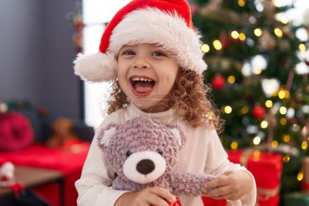 Photo for Adorable blonde girl hugging teddy bear standing by christmas tree at home - Royalty Free Image