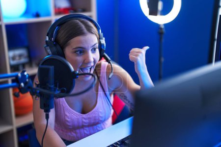Téléchargez les photos : Jeune femme blonde jouant à des jeux vidéo parlant avec un microphone avec le public pointant le pouce vers le côté souriant heureux avec la bouche ouverte - en image libre de droit