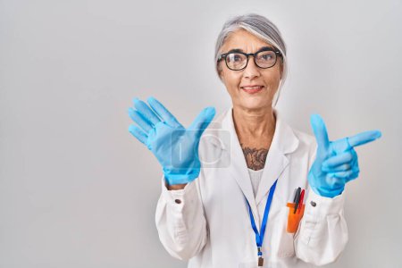 Foto de Mujer de mediana edad con el pelo gris vistiendo túnica científica mostrando y señalando hacia arriba con los dedos número siete mientras sonríe confiado y feliz. - Imagen libre de derechos