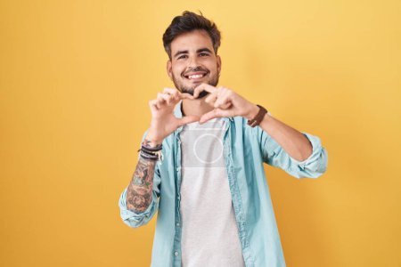 Photo for Young hispanic man with tattoos standing over yellow background smiling in love doing heart symbol shape with hands. romantic concept. - Royalty Free Image