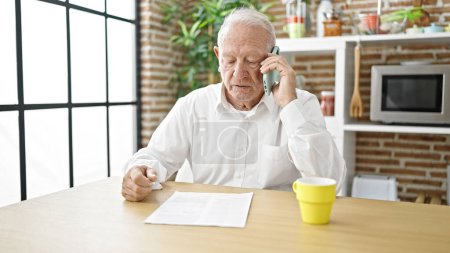 Foto de Senior hombre de pelo gris enojado discutiendo en el teléfono inteligente sentado en la mesa en el comedor - Imagen libre de derechos