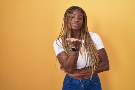 Photo for African american woman with braided hair standing over yellow background looking at the camera blowing a kiss with hand on air being lovely and sexy. love expression. - Royalty Free Image