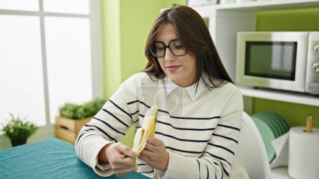 Photo for Young beautiful hispanic woman eating banana sitting on table at dinning room - Royalty Free Image