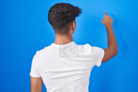 Photo for Hispanic teenager standing over blue background posing backwards pointing ahead with finger hand - Royalty Free Image