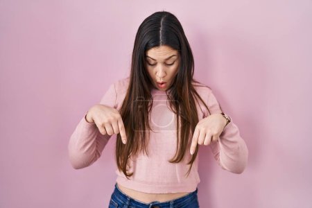 Téléchargez les photos : Jeune femme brune debout sur fond rose pointant vers le bas avec les doigts montrant la publicité, visage surpris et bouche ouverte - en image libre de droit