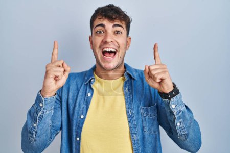 Téléchargez les photos : Jeune homme hispanique debout sur fond bleu souriant étonné et surpris et pointant vers le haut avec les doigts et les bras levés. - en image libre de droit