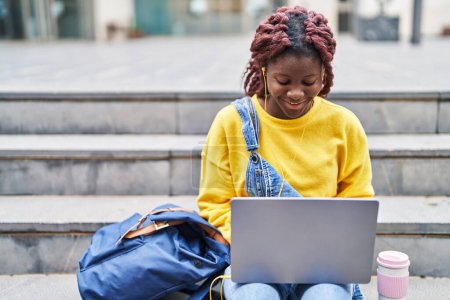Téléchargez les photos : Étudiante afro-américaine utilisant un ordinateur portable et des écouteurs assis sur les escaliers à l'université - en image libre de droit