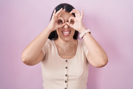 Téléchargez les photos : Jeune femme hispanique debout sur fond rose faisant un geste correct comme des jumelles sortant la langue, les yeux regardant à travers les doigts. expression folle. - en image libre de droit