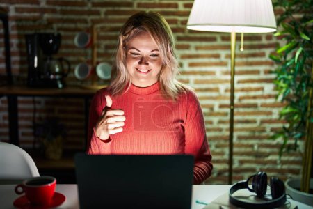 Téléchargez les photos : Femme blonde utilisant un ordinateur portable la nuit à la maison faisant des pouces heureux geste avec la main. approuvant l'expression regardant la caméra montrant le succès. - en image libre de droit