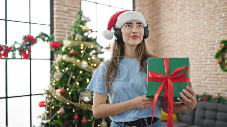 Foto de Joven mujer hispana hermosa escuchando música sosteniendo regalo celebrando la Navidad en casa - Imagen libre de derechos