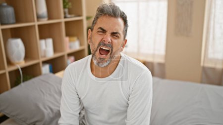 Foto de Hombre hispano joven agotado con el pelo gris, bostezando boca abierta mientras está sentado en la cama acogedora, luchando para despertar en el dormitorio - Imagen libre de derechos