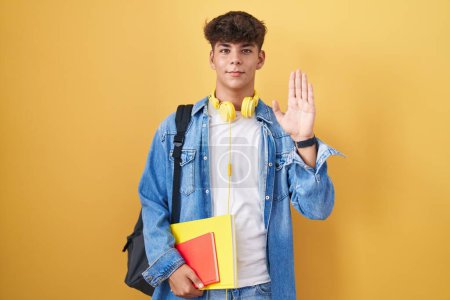 Téléchargez les photos : Adolescent hispanique portant un sac à dos d'étudiant et tenant des livres renonçant à dire bonjour heureux et souriant, geste de bienvenue amical - en image libre de droit