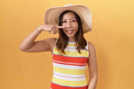 Foto de Mujer china de mediana edad con sombrero de verano sobre fondo amarillo señalando con el dedo de la mano a la cara y la nariz, sonriendo alegre. concepto de belleza - Imagen libre de derechos
