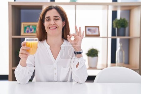 Téléchargez les photos : Brunette femme boire un verre de jus d'orange souriant positif faire ok signe avec la main et les doigts. expression réussie. - en image libre de droit