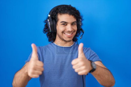 Photo for Hispanic man with curly hair listening to music using headphones approving doing positive gesture with hand, thumbs up smiling and happy for success. winner gesture. - Royalty Free Image