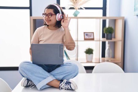 Téléchargez les photos : Jeune femme hispanique utilisant un ordinateur portable assis sur la table portant des écouteurs souriant avec la main sur l'oreille en écoutant une rumeur ou des commérages. concept de surdité. - en image libre de droit