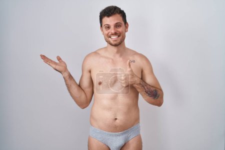 Foto de Joven hombre hispano de pie sin camisa usando ropa interior que muestra la palma de la mano y haciendo buen gesto con los pulgares hacia arriba, sonriendo feliz y alegre - Imagen libre de derechos