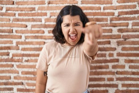 Foto de Mujer hispana joven de pie sobre la pared de ladrillos señalando disgustado y frustrado a la cámara, enojado y furioso con usted - Imagen libre de derechos