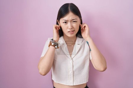 Téléchargez les photos : Jeune femme chinoise debout sur fond rose couvrant les oreilles avec des doigts avec une expression agacée pour le bruit de la musique forte. concept sourd. - en image libre de droit