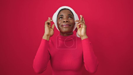 Photo for African american woman smiling confident wearing christmas hat crossing fingers for luck over isolated red background - Royalty Free Image