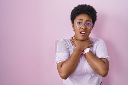 Foto de Mujer afroamericana joven de pie sobre fondo rosa gritando y sofocar porque estrangulamiento doloroso. problema de salud. concepto de asfixia y suicidio. - Imagen libre de derechos