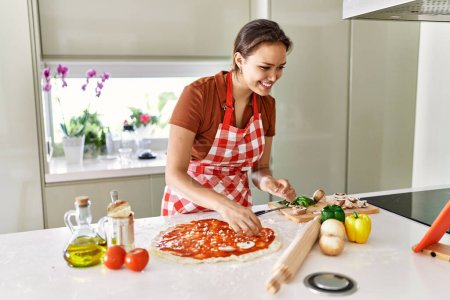 Foto de Joven hermosa mujer hispana preparando pizza italiana viendo receta en línea en touchpad en la cocina - Imagen libre de derechos