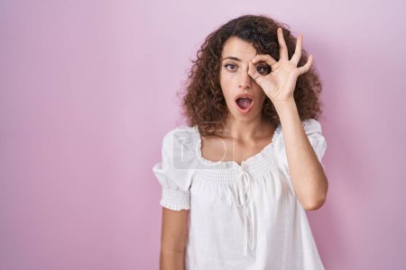 Téléchargez les photos : Femme hispanique aux cheveux bouclés debout sur fond rose faisant un geste correct choqué par le visage surpris, les yeux regardant à travers les doigts. expression incrédule. - en image libre de droit
