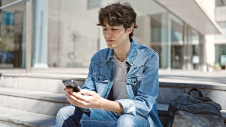 Téléchargez les photos : Jeune homme hispanique étudiant utilisant smartphone assis sur les escaliers à l'université - en image libre de droit